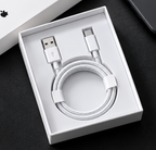 A white braided USB-A to USB-C charging cable neatly coiled inside an open white retail box, photographed on a dark surface with part of a box lid and a phone visible in the background.