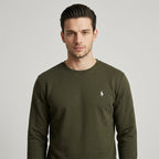 A man wearing an olive green long-sleeve crewneck sweatshirt with the white embroidered Polo Ralph Lauren pony logo on the left chest, paired with blue jeans against a plain white studio background.