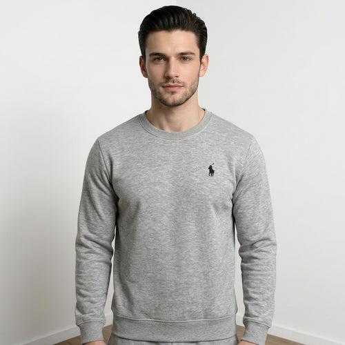 A man wearing a heather gray long-sleeve crewneck sweatshirt with a small black embroidered polo player logo on the left chest, set against a plain light gray wall.