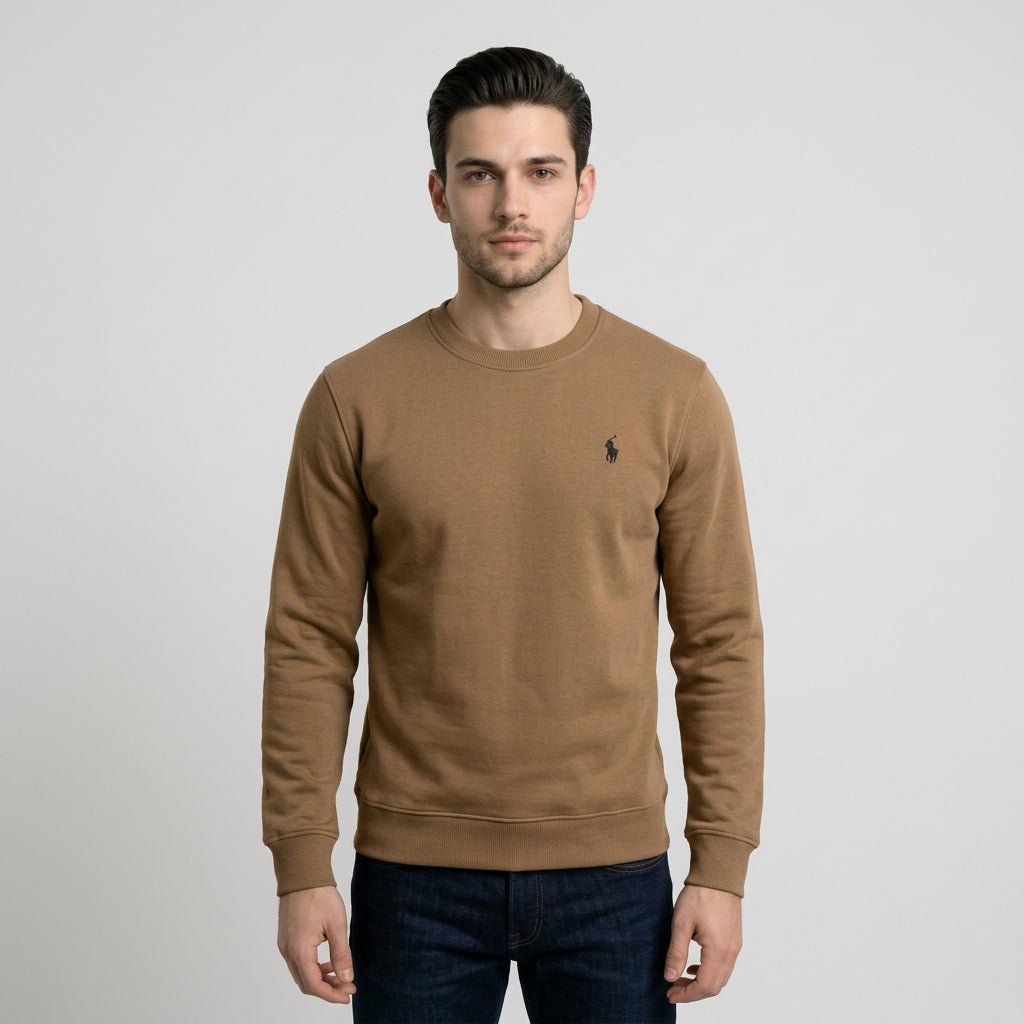 A man wearing a tan crewneck sweatshirt with a small black Polo Ralph Lauren pony logo on the left chest, photographed against a light gray studio background.