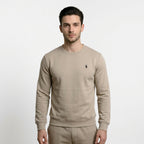 A man wearing a beige long-sleeve crewneck sweatshirt by Polo Ralph Lauren, featuring the brand's signature polo player logo on the left chest and a hanging purple brand tag.