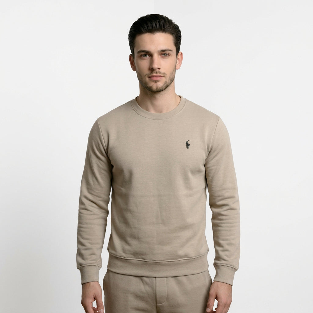 A man wearing a beige long-sleeve crewneck sweatshirt by Polo Ralph Lauren, featuring the brand's signature polo player logo on the left chest and a hanging purple brand tag.